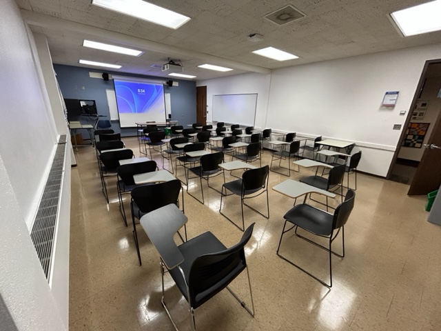Front of the classroom with student desks arranged in rows. A large white board and projector screen is at the front.