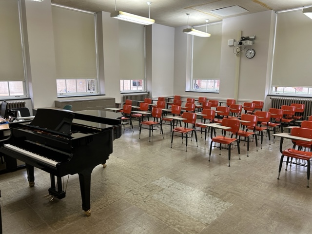 Back of the classroom with large windows on the walls. A grand piano is in the front with student desks behind it.