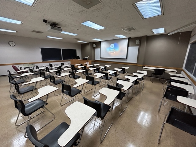 Front of the classroom with student desks in rows, a large whiteboard and projector screen.