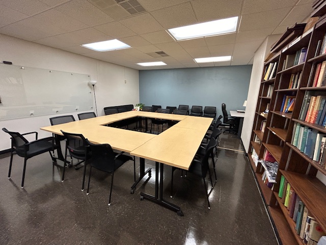 Front of the conference room with tables and chairs in a circular formation, a whiteboard, and lots of bookshelves are on the right wall.