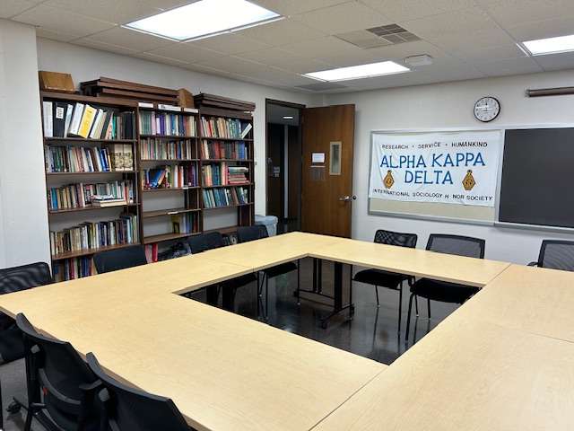 Front of the conference room with tables and chairs in a circular formation, a black board, and lots of bookshelves are on the left wall.