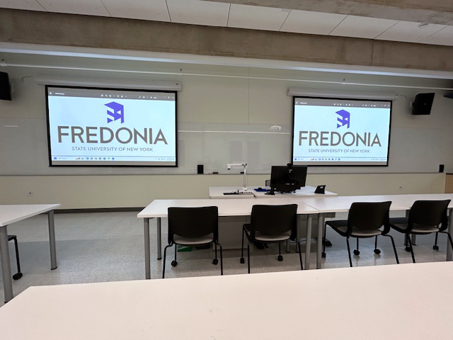Front of the classroom with a large white board and two projector screens.