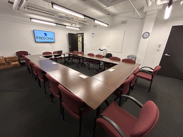 Front of the conference room with several tables and chairs and a large tv